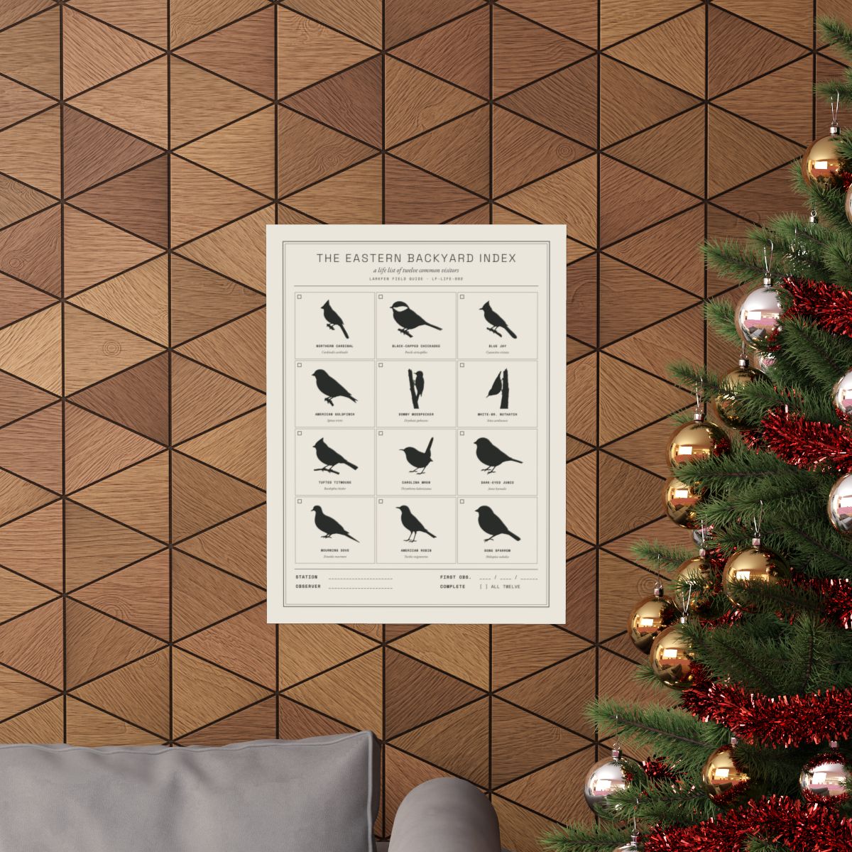 The Eastern Backyard Index poster framed on a warm wood-tile wall in a lived-in room — twelve common eastern visitors drawn in silhouette in a hand-set grid, with a checkbox per species and a station and observer line.